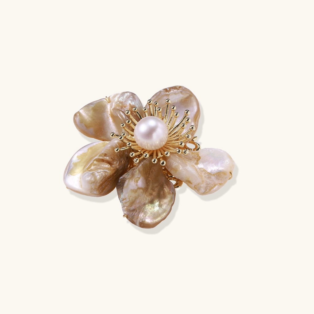 Elegant floral pin with peachy shell petals, gold radiating spokes, and white freshwater pearl center on cream background.