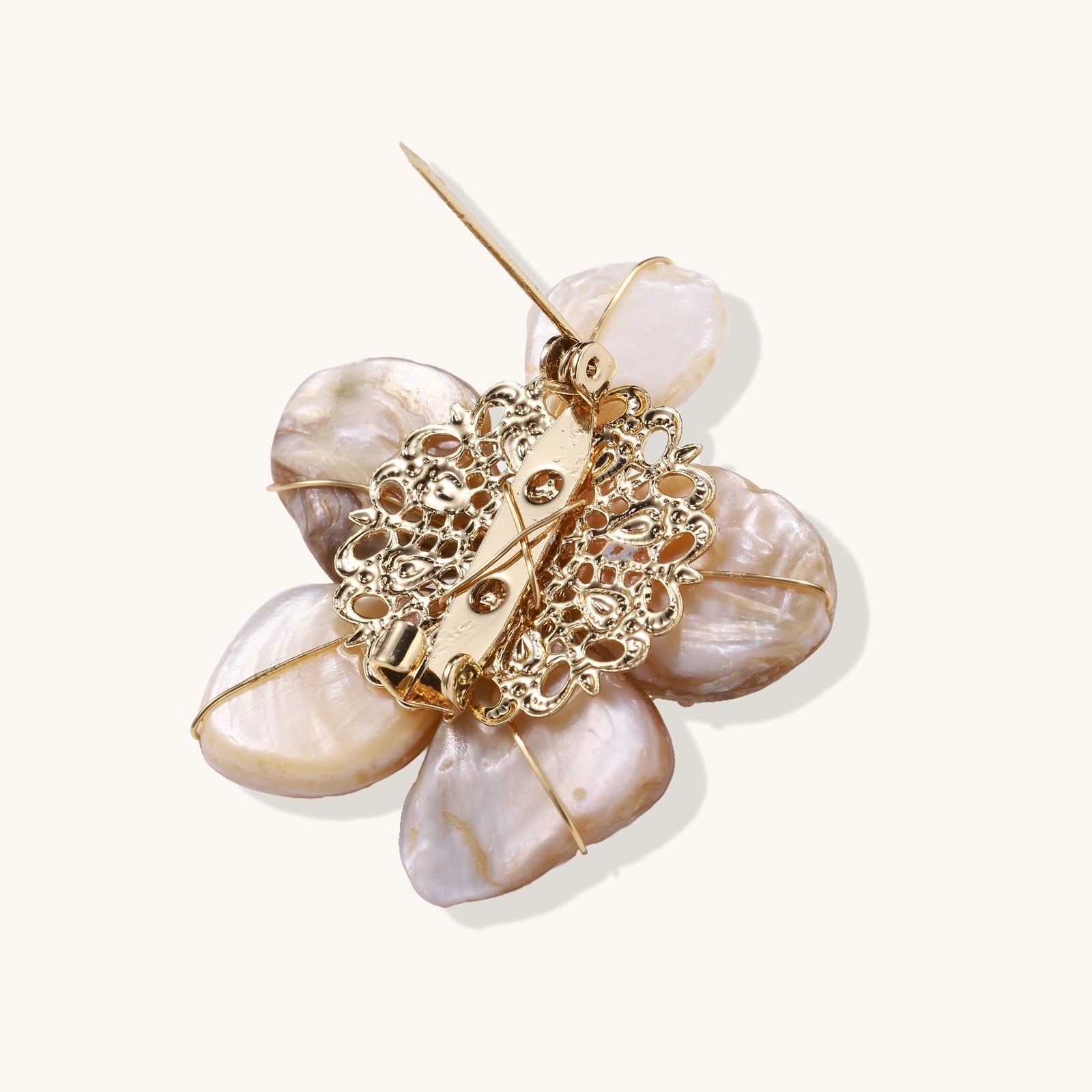 Elegant floral pin with peachy shell petals, gold radiating spokes, and white freshwater pearl center on cream background.