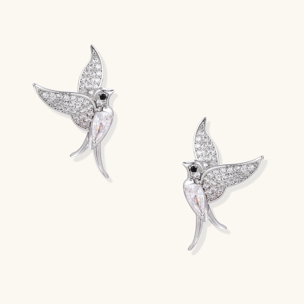 Pair of gold-plated hummingbird earrings with crystal pavé wings and black stone eyes on neutral background.