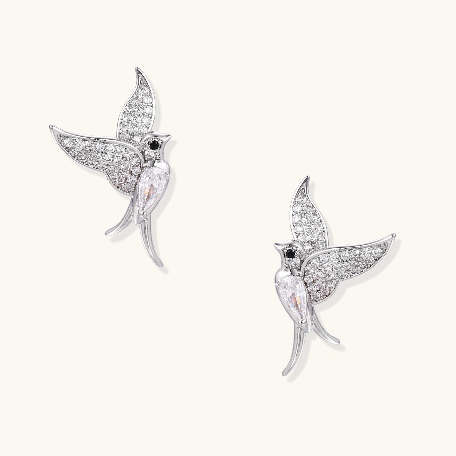 Pair of gold-plated hummingbird earrings with crystal pavé wings and black stone eyes on neutral background.