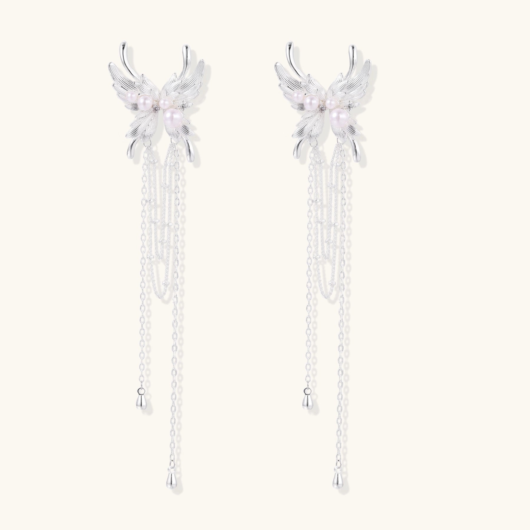 Elegant silver-plated drop earrings with wing-shaped tops, white pearl accents, and long chain tassels on white background.