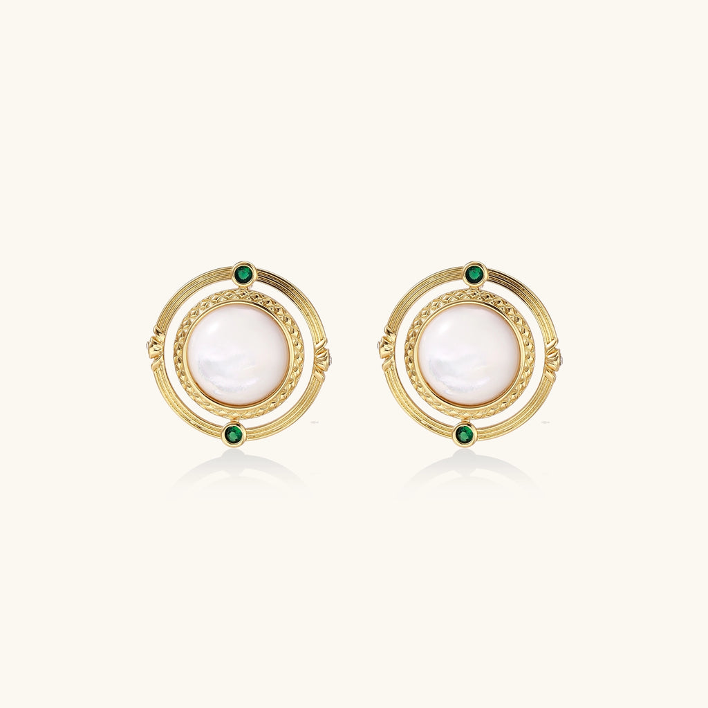 Close-up of gold-plated circular earrings with white mother-of-pearl centers and green zirconia accents on cream background.