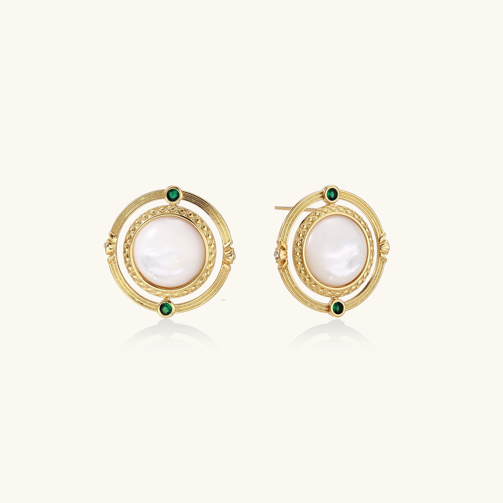 Close-up of gold-plated circular earrings with white mother-of-pearl centers and green zirconia accents on cream background.