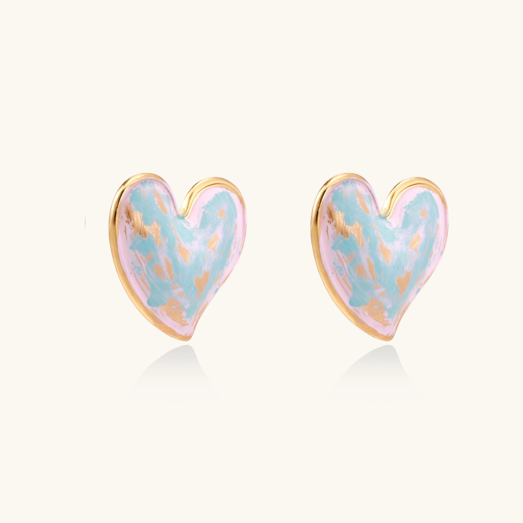 Heart-shaped stud earrings with iridescent blue-pink enamel finish and gold-plated brass frames against cream background