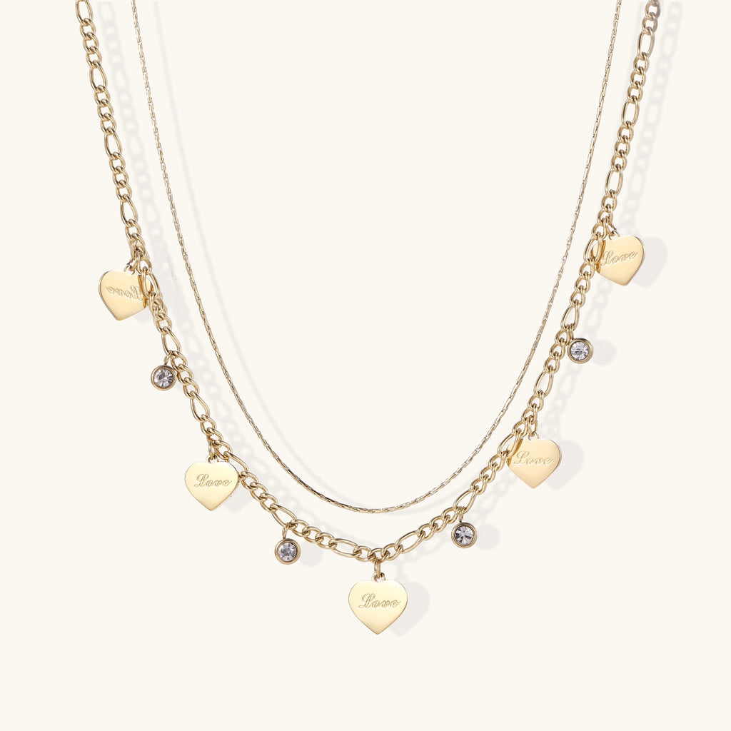 Two layered gold-plated necklaces with heart pendants engraved 'Love' and sparkling crystal charms on white background.