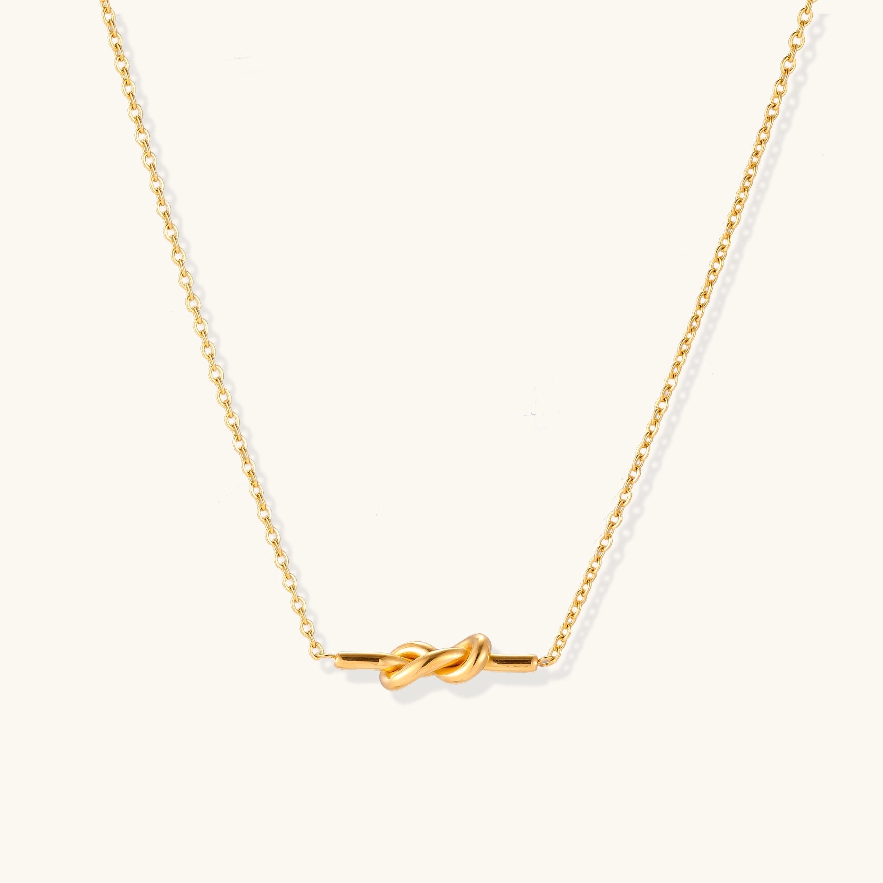 Dainty 18K gold plated stainless steel chain necklace with minimalist knot pendant on cream background.
