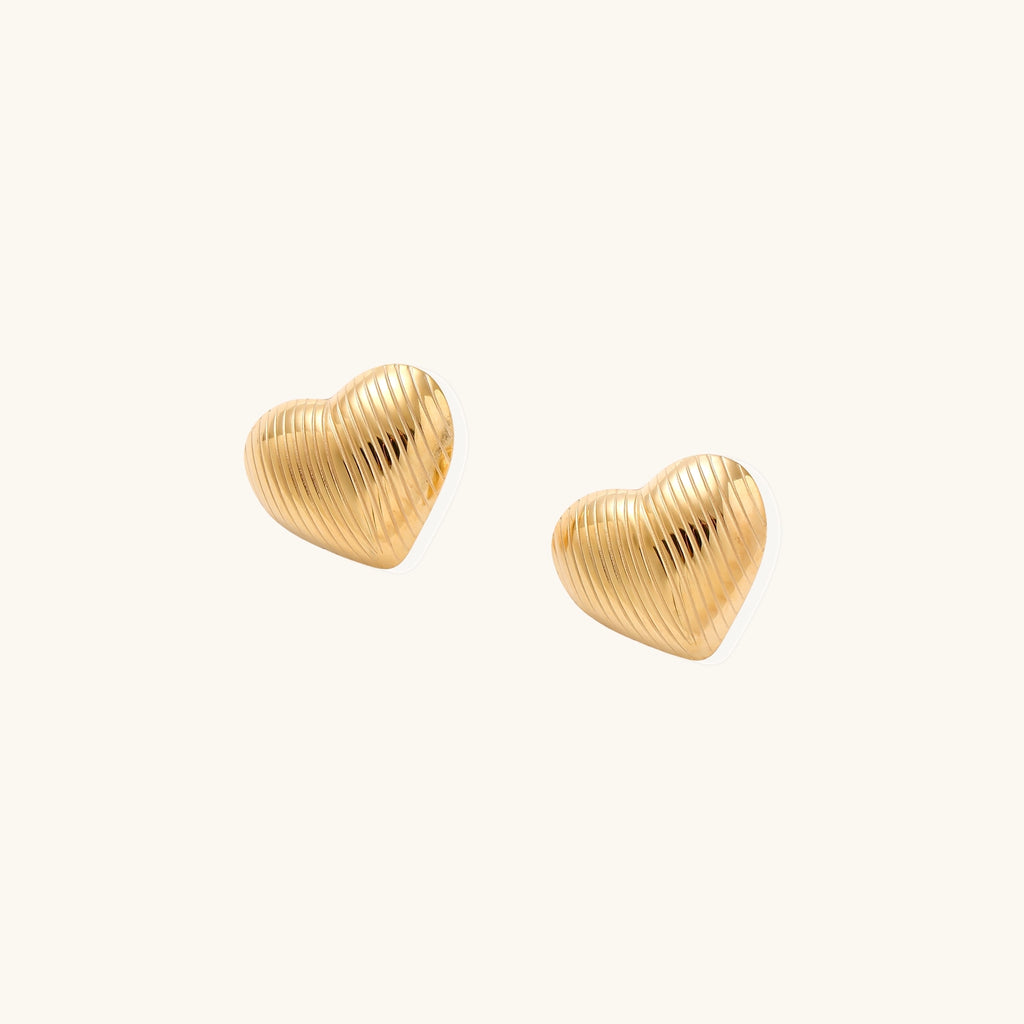 Close-up of ridged heart-shaped gold-plated stud earrings with textured surface pattern on cream background.