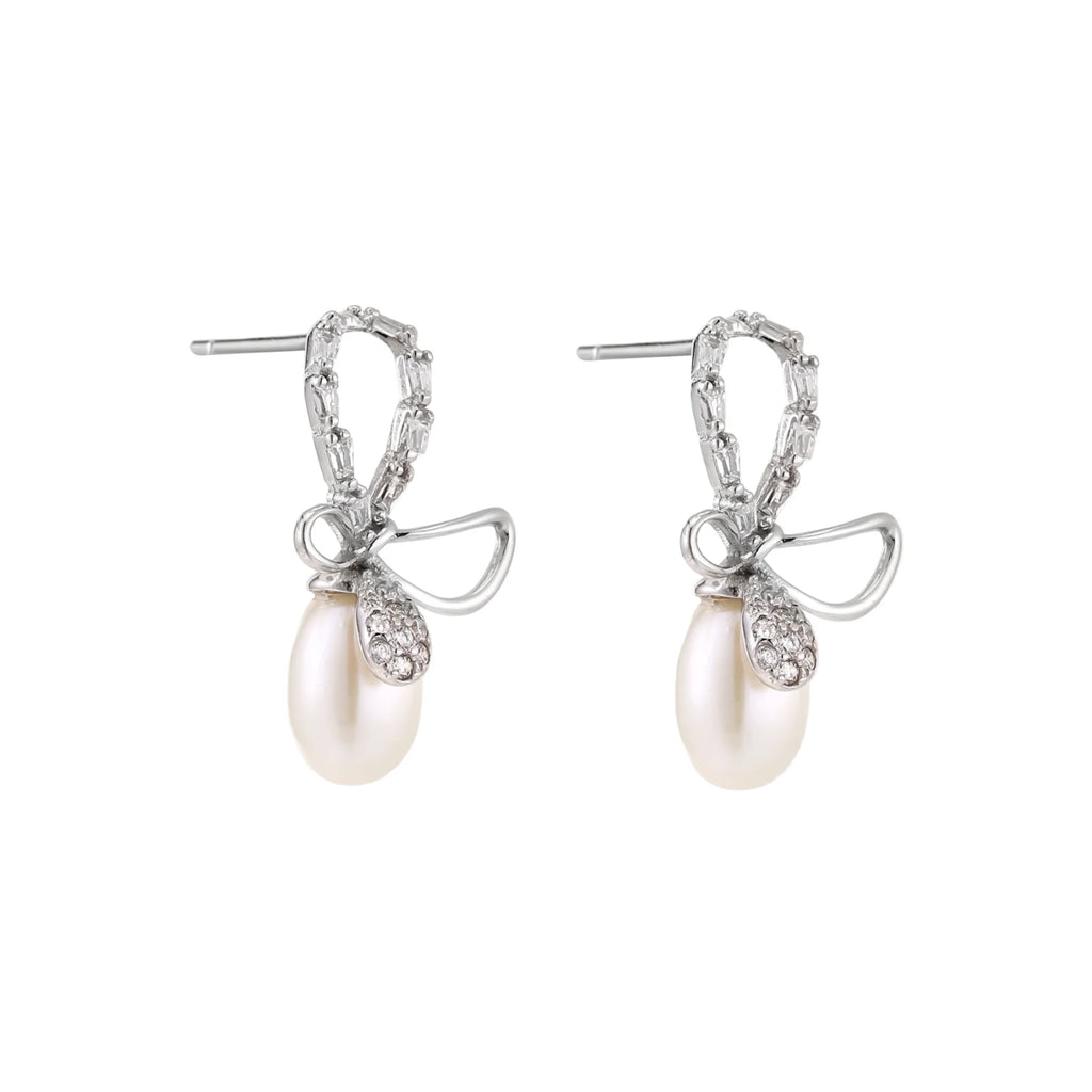Silver bow-shaped stud earrings with baguette crystals and dangling white freshwater pearls on cream background